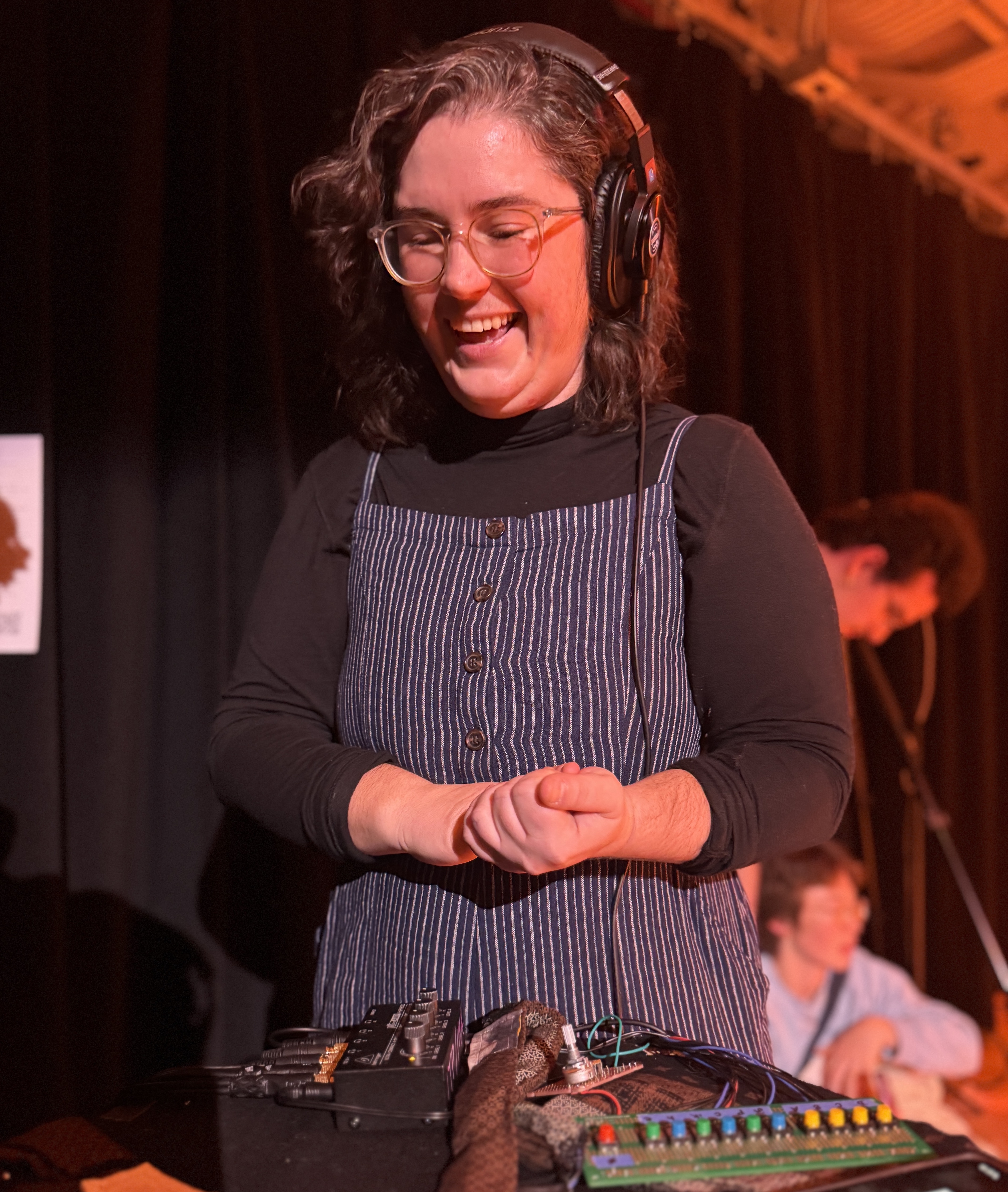 devan (me), a white person with glasses and curly brown hair wearing headphones, hands clasped and grinning excitedly down at my textiles and circuitry project 'an acchording'
